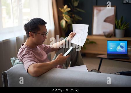 Asian young adult man sitting on sofa holding smartphone and examining financial documents, laptop with charts on table in background, working remotely in modern living room Stock Photo