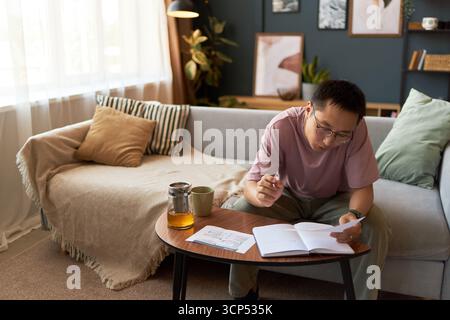 Young adult Asian man sitting at coffee table reading notebook and holding pen, studying or working at home with documents and tea, modern living room with sofa and decor in background Stock Photo