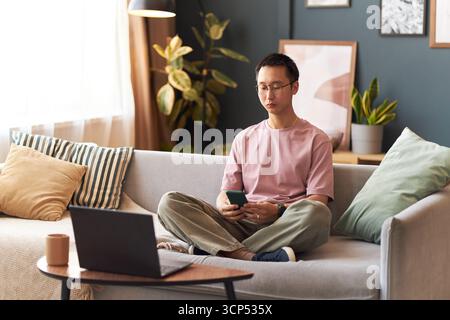 Asian young adult man sitting cross legged on sofa using smartphone while looking at laptop screen in modern living room, appearing focused and engaged with digital devices Stock Photo