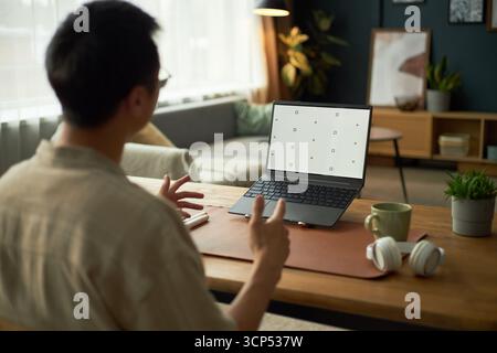 Asian young adult man gesturing while sitting at desk in home office, interacting with laptop displaying calibration screen, wireless headphones and coffee mug on table Stock Photo