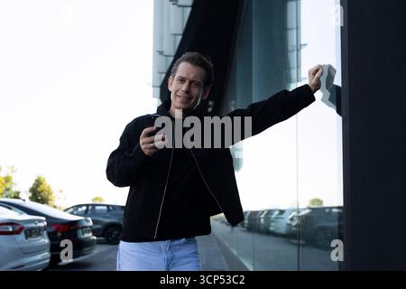 Smiling man stands outside a modern building while checking his smartphone in bright daylight Stock Photo
