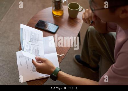 Young adult Asian man sitting at table analyzing financial documents with pen in hand, smartphone and notebook nearby, focusing on paperwork, partial face visible Stock Photo