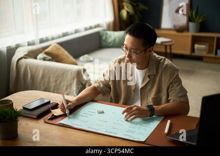 Asian young adult man sitting at desk writing on large sheet of paper with pen, working on project or brainstorming ideas, smartphone and laptop placed nearby on wooden table Stock Photo