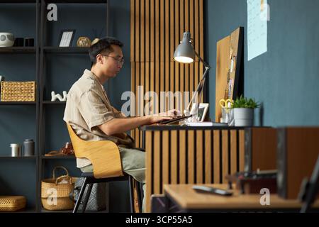 Asian young adult man working at desk using laptop in modern home office, sitting on chair with focused expression, hands typing on keyboard, shelves and lamp in background Stock Photo