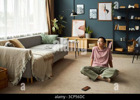 Asian young adult man sitting cross legged on carpeted floor in modern living room looking upward with thoughtful expression, digital tablet lying nearby on floor Stock Photo