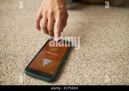 Young adult Caucasian man hand interacting with smartphone lying on carpet, finger touching screen displaying meditation app session interface, partial body visible Stock Photo