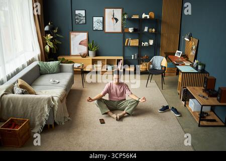 Asian young adult man meditating cross legged on carpet in modern living room, closing eyes and practicing mindfulness with smartphone placed in front, surrounded by home decor Stock Photo