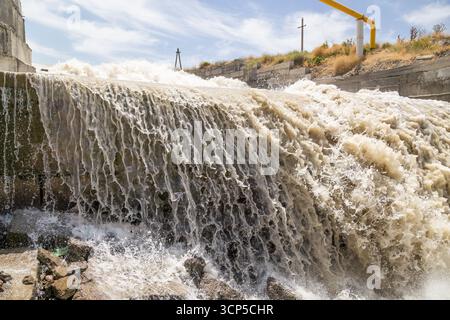 Water cascades forcefully over a dam, creating a turbulent scene in a rural outdoor setting. Daylight enhances the dynamic flow and intensity of the w Stock Photo