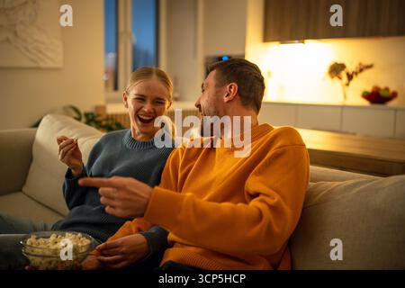 Smiling young couple enjoying popcorn watching movie together on living room sofa, family night Stock Photo