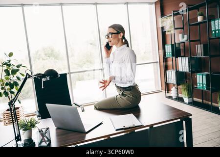 Confident businesswoman speaking on the phone while working in a modern office environment, surrounded by professional elements Stock Photo