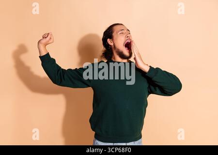 Young man in a green sweatshirt yawning with a raised arm against a beige background, expressing tiredness Stock Photo