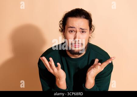 Young man with a confused facial expression gesturing with hands against a beige background, wearing a green sweatshirt Stock Photo