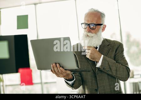 Professional elder businessman analyzing data on a laptop in a modern office environment with a thoughtful expression Stock Photo