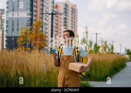 Smiling courier delivering a parcel in a city, using a mobile app for navigation, showcasing modern logistics, e-commerce, and the gig economy, providing efficient last-mile service Stock Photo