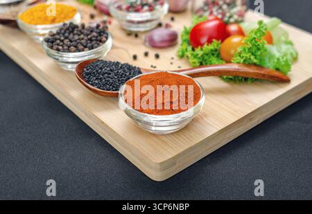 Bright paprika in small transparent bowl close-up. Paprika among vegetables, spices and seasonings on wooden board. Black tablecloth. Culinary inspira Stock Photo