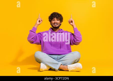 Funny man in purple hoodie seated on yellow background pointing upwards with enthusiasm, expressing excitement Stock Photo