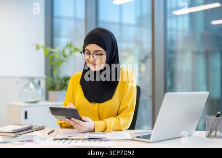 Muslim businesswoman wearing a black hijab and glasses, working in a modern office, concentrating while using a calculator for financial planning and accounting tasks on her desk Stock Photo