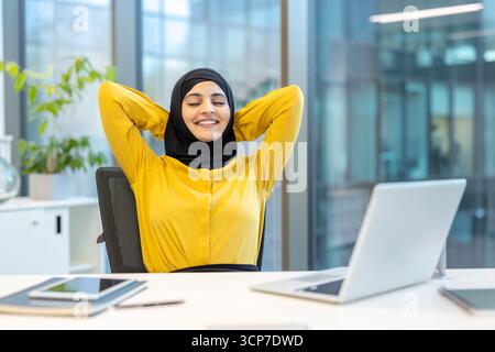 Muslim businesswoman is stretching and relaxing in her office chair, feeling great and happy with closed eyes and hands behind her head after a successful day of work Stock Photo