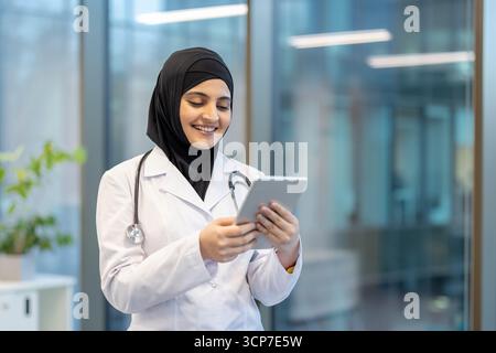 Young arabian female doctor in hijab and white coat, smiling while using a tablet to review electronic health records and manage patient data in a modern clinic setting Stock Photo