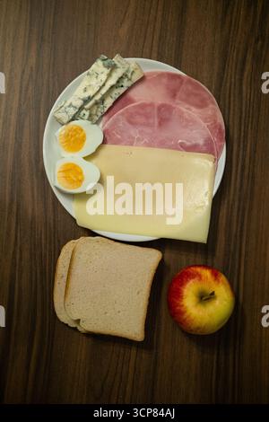 Breakfast plate with ham, cheese, boiled egg, bread and apple on a wooden table, illustrating healthy morning meal and nutrition concept. Stock Photo