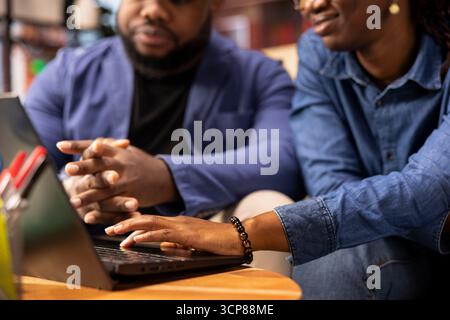 Casual man and woman opening laptop on the coffee table to solve daily tasks, doing remote freelance work online. Black couple surfing the web and multitasking in their loft apartment. Close up. Stock Photo