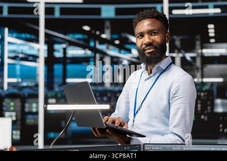 Portrait of cheerful data center engineer monitoring system performance with laptop, identifying bottlenecks. Upbeat man in server hub troubleshooting errors, implementing optimizations Stock Photo