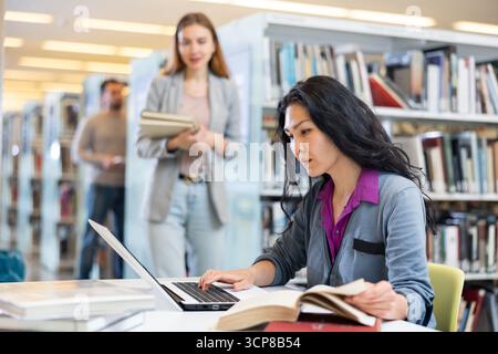 Asian woman student in the library, searches for information in books and types it in a laptop Stock Photo