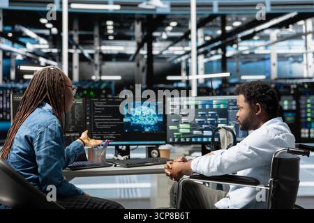 Person with paraplegia in inclusive server room and coworker using artificial intelligence to reduce downtime. Engineer with disability in wheelchair and colleague implement AI in data center Stock Photo