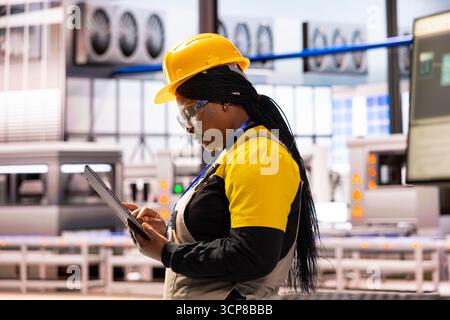 Focused factory manager holding tablet, monitoring production operations in industrial workspace. Black female software specialist making system upgrades and ensuring efficient workflow in warehouse. Stock Photo