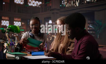 Multicultural students group discussing a financial project for a university course, using laptop and textbooks with class notes in the library. Entrepreneurship and management skills. Camera B. Stock Photo