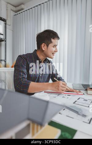 Young male architect working at office desk, drawing construction plans and using drafting tools, focused on building design while showing creativity Stock Photo