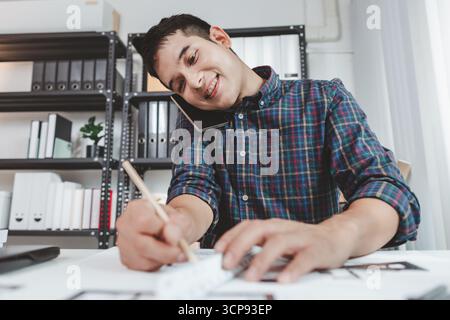 Young male architect working at office desk, drawing construction plans and using drafting tools, focused on building design while showing creativity Stock Photo