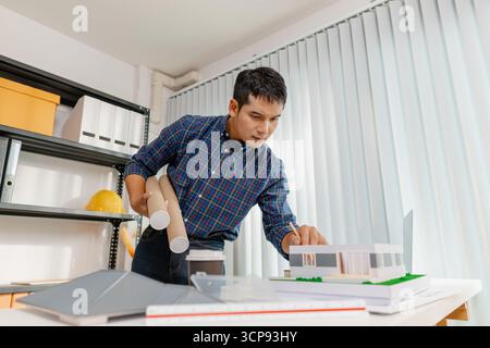 A young architect works on house design plans at his desk, focusing on drawings and scale models. The scene conveys creativity, dedication, and precis Stock Photo