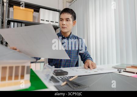 Young male architect working thoughtfully at office desk, analyzing house blueprints and construction plans, reviewing details carefully to ensure acc Stock Photo