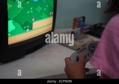 A nostalgic image of a vintage Super Nintendo (SNES) and controller on a desk. A girl plays a classic game on an old CRT TV, capturing 90s gaming. Stock Photo