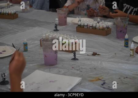 Kids painting small figures at a workshop. The table is covered in plastic, with paints and water cups visible. Stock Photo