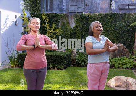Female friends practicing yoga prayer pose in backyard with fitness smartwatch and hedge Stock Photo