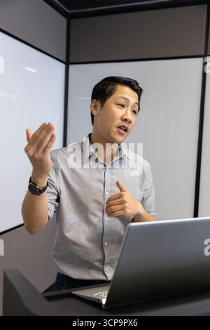 Asian man presenting behind podium in meeting room with laptop computer, frosted glass panels Stock Photo