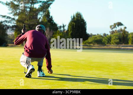 African American man with cap crouching on putting green placing golf ball with putter, copy space Stock Photo