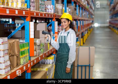 Female warehouse worker using handheld scanner scanning boxes on orange-blue racks in aisle by cart Stock Photo