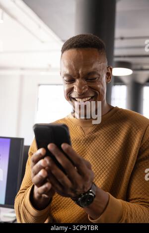 African american man checking smartphone at desk in modern office, wearing wristwatch Stock Photo