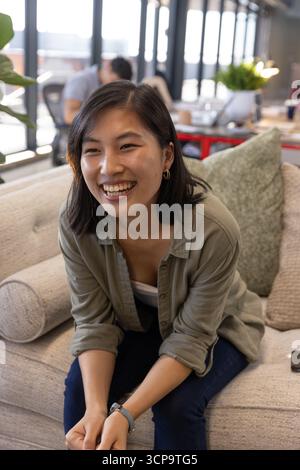 Diverse coworkers laughing, sharing ideas on grey sofa in office lounge with potted plants Stock Photo