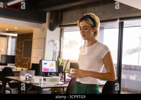 Woman standing in office workspace with ceramic mug reviewing computer monitors, copy space Stock Photo