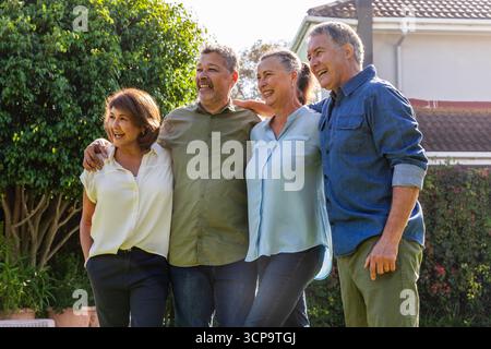 Senior friends standing arm-in-arm in backyard under tree by roof house with hedge Stock Photo