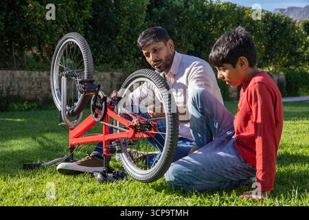 Indian father with son kneeling on suburban lawn repairing red bicycle using wrench and toolkit Stock Photo