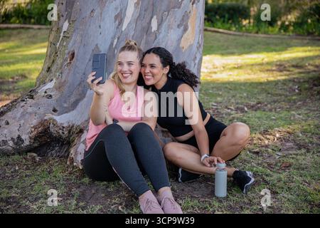Diverse female friends sitting by tree trunk in park in sportswear taking selfie using smartphone Stock Photo