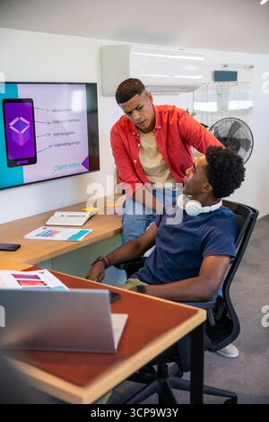Diverse male coworkers collaborating on mobile app prototype at office desk with laptop and monitor Stock Photo