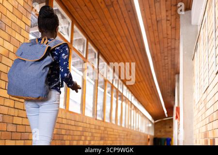 African American child walking down school hall with denim backpack near windows and recycling bins Stock Photo
