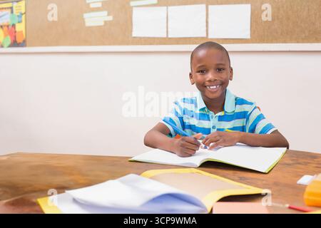 African American boy smiling at camera while holding pencil over open notebook at classroom desk Stock Photo