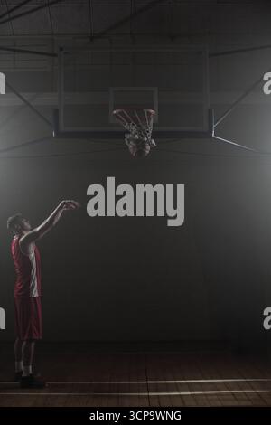 Basketball falling through net on wooden gym court under spotlights with deep shadows Stock Photo